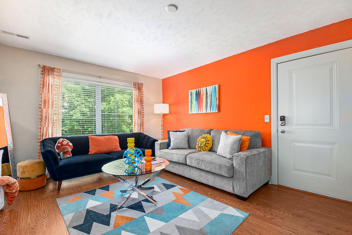 A bright living area features an orange accent wall and large windows allowing natural light. Seating includes a dark couch and a light gray sofa. A round glass coffee table is center stage, complemented by a colorful patterned rug, creating a cheerful and inviting space.