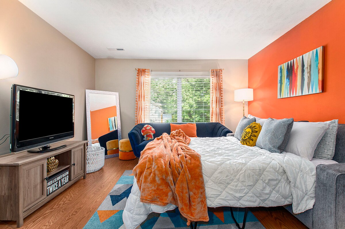 A cozy bedroom features a spacious bed draped with a light quilt. An orange accent wall contrasts with other soft hues. Natural light filters through large windows covered by sheer drapes. A large TV is positioned on a wooden stand next to a decorative mirror, providing a vibrant atmosphere.