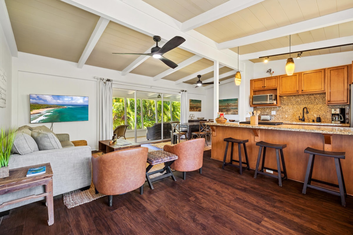 An open-concept living area features a light-toned sofa and two leather chairs facing a wall-mounted TV. The kitchen, with wooden cabinetry and modern appliances, includes a breakfast bar with matching stools. Large windows invite natural light and offer views of lush greenery.