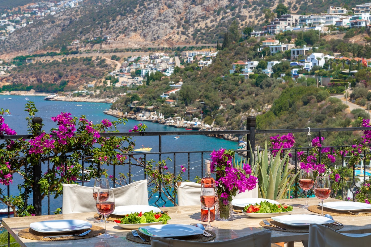 A dining table is set outdoors with plates, glasses, and fresh salads, overlooking the sea. Bougainvillea flowers in vibrant purple frame the view, which includes the sparkling water and hills dotted with villas, creating a scenic and inviting dining environment.
