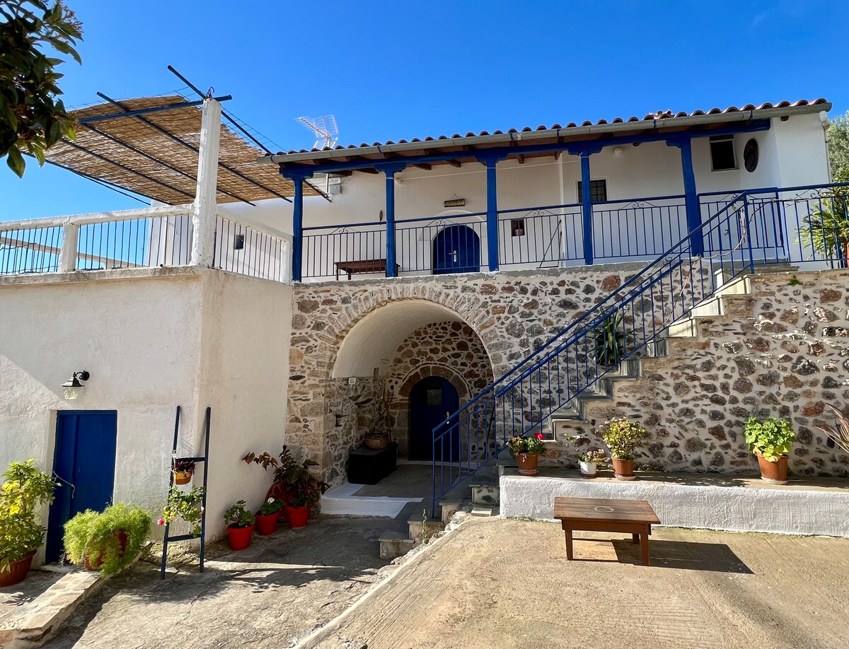A traditional stone house features a two-story layout, with a balcony supported by blue railings. An exterior staircase leads to the entrance, flanked by potted plants. The ground level is adorned with a seating area under a shaded pergola, enhancing the connection with the outdoor surroundings.