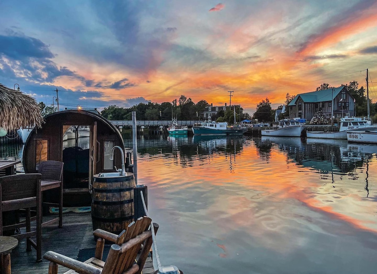 A serene marina scene is presented at sunset, with vibrant hues of orange and pink reflecting on the calm water. Various boats are anchored in the background, while the foreground features rustic seating made from wood and a barrel sink.