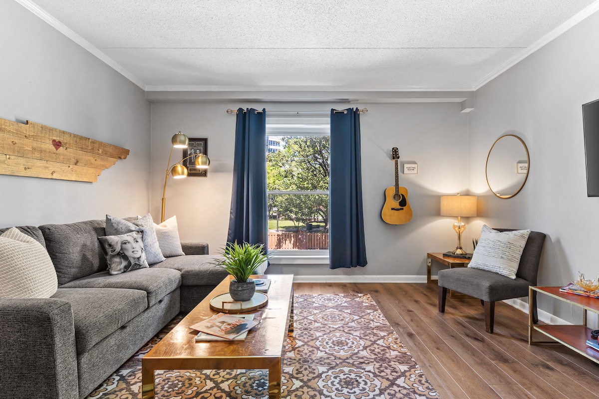 The inviting living area features a comfortable gray sofa with decorative pillows and a wooden coffee table. A warm light illuminates the space, complemented by a standing lamp. An acoustic guitar is displayed on the wall, with a window allowing natural light and views of greenery outside.