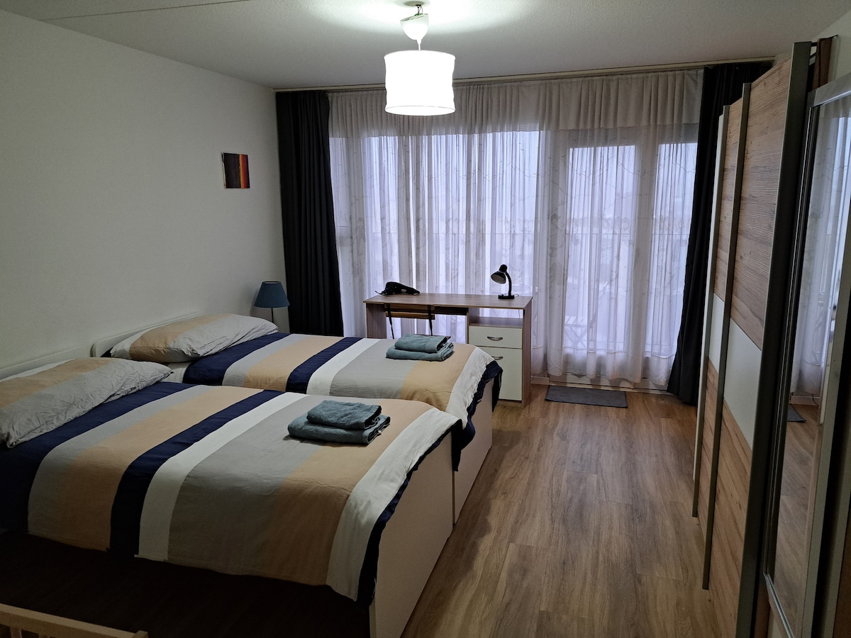 A spacious bedroom features two beds with striped linens. Natural light filters through floor-to-ceiling sheer curtains, illuminating the wood-toned flooring. A desk with a lamp and a small bedside table complete the functional layout, providing a comfortable space for relaxation or work.