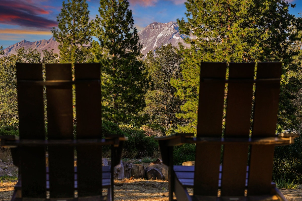 Two Adirondack chairs are positioned to face a stunning view of majestic mountains. Lush trees frame the scene, and a colorful sky transitions from sunset to twilight, creating a serene outdoor setting for relaxation.
