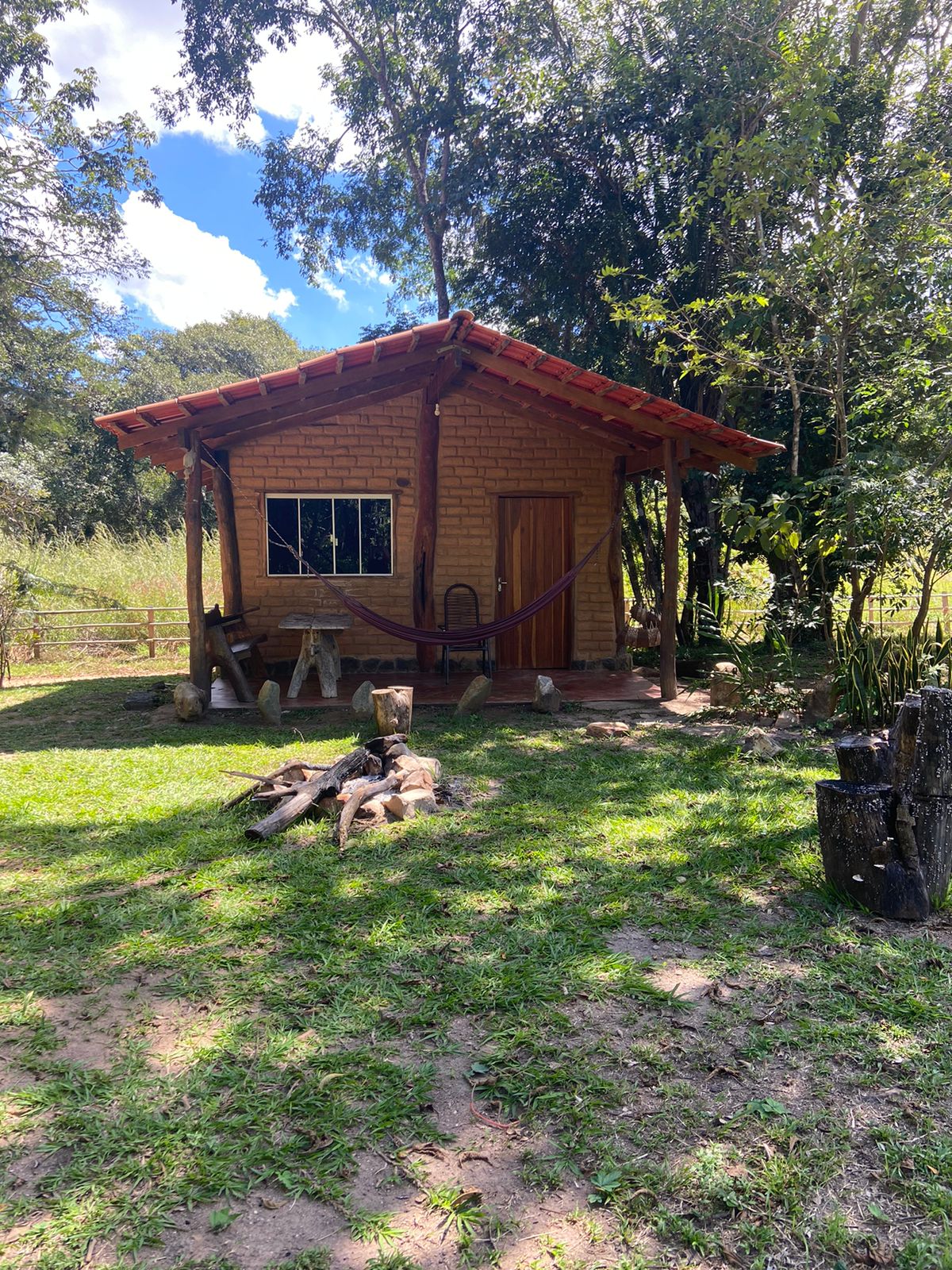 A rustic cabin constructed from natural materials is surrounded by greenery. The structure features a small window, a wooden door, and a hammock hanging outside. A fire pit made of logs is set in the grassy area in front of the cabin.