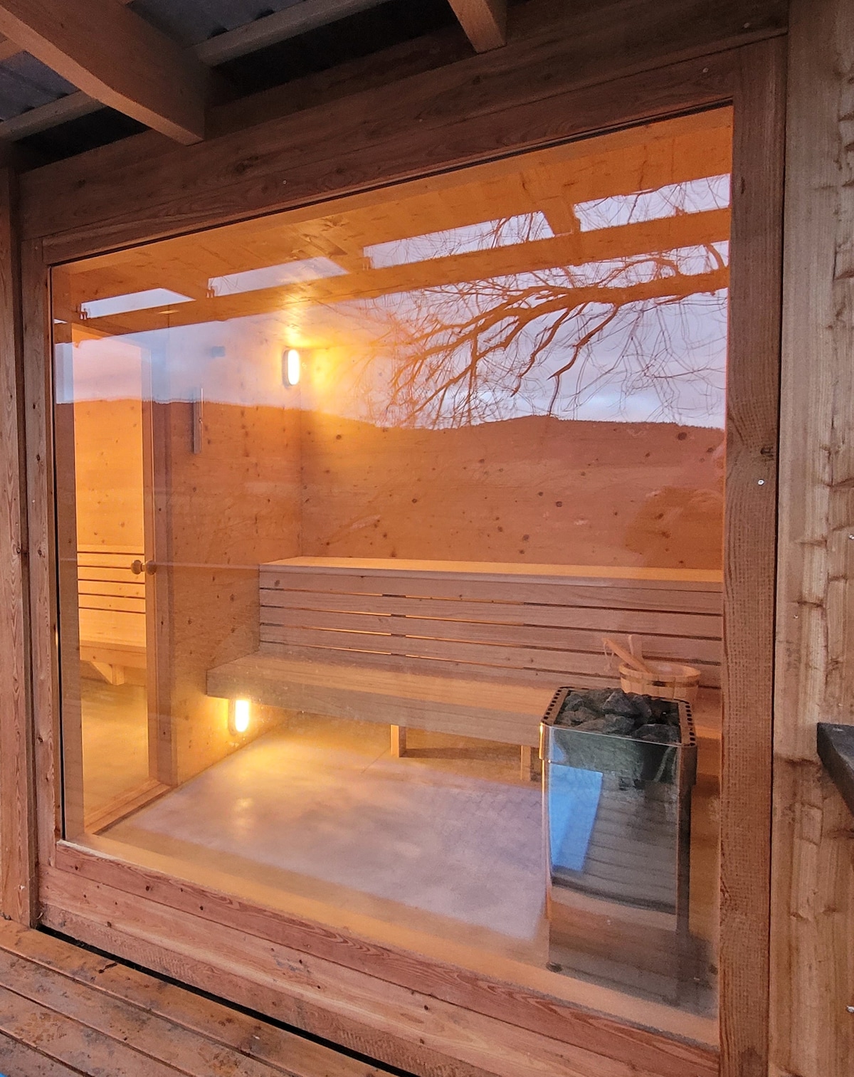 An inviting wooden sauna is visible through large glass panels, showcasing a minimalistic interior with wooden benches. Soft lighting enhances the tranquil atmosphere. A small metal stove is positioned near the seating area, providing warmth. The structure is surrounded by natural scenery, promoting relaxation.