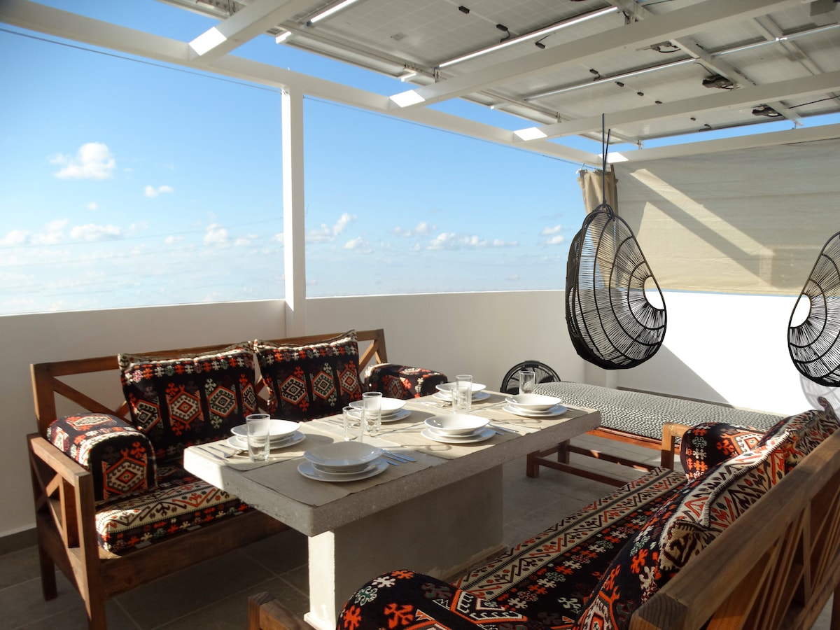 A rooftop seating area features a large concrete table surrounded by patterned seating. Two hanging chairs are positioned for relaxation. The space is illuminated by natural light, with a clear sky visible in the background, providing a serene atmosphere for gatherings.