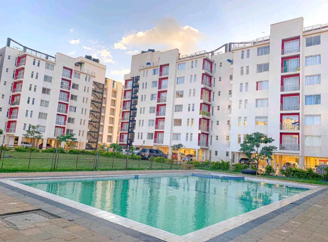 A clear view of the swimming pool reflects the surrounding apartment buildings, which feature a combination of white and red facades. The landscaped area includes a grassy lawn and decorative plants, providing a serene environment in the outdoor space.