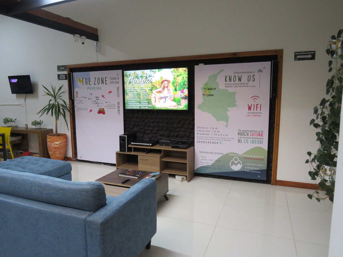 A welcoming common area is highlighted by a comfortable blue sofa and a modern media console. Informative banners decorate the walls, and a large screen displays various amenities. Natural light fills the space, creating an inviting atmosphere for guests to relax or socialize.