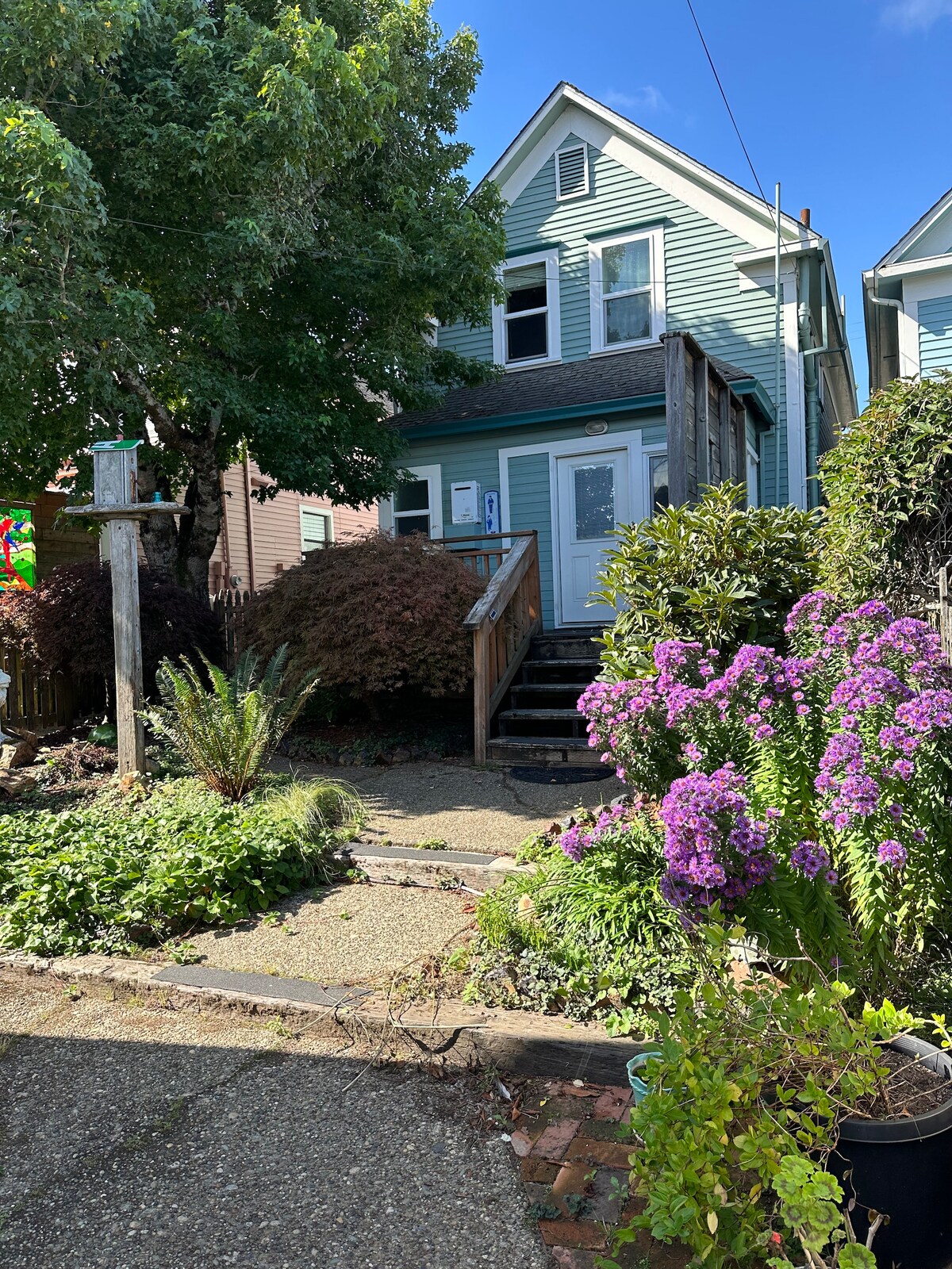 A charming two-story house is surrounded by lush greenery and vibrant purple flowers. A stone pathway leads to a wooden staircase that ascends to a welcoming entrance. The exterior is painted in soft blue hues, contributing to a cozy appearance in the neighborhood.
