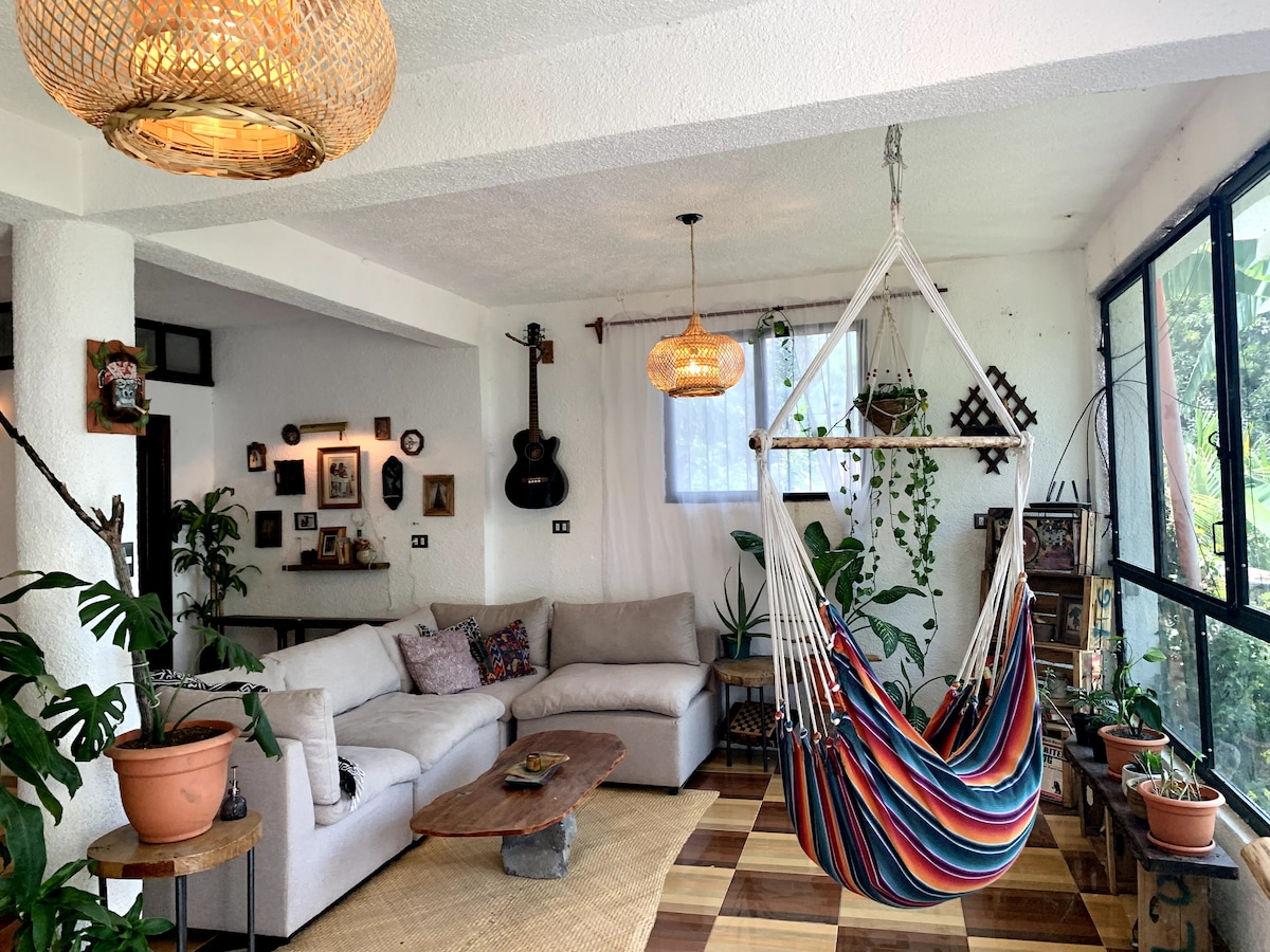A bright living area features a comfortable sofa and a colorful hammock hanging from the ceiling. Natural light filters through large windows, enhancing the space. Potted plants add a touch of greenery, while decorative wall art and a guitar create a welcoming ambiance.