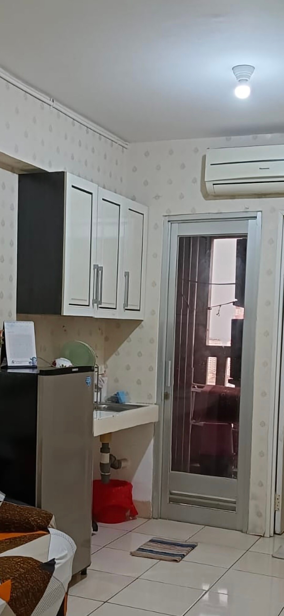 The image shows a modern kitchenette area with upper cabinets and a small sink. A compact refrigerator is positioned to the left. An air conditioning unit is mounted on the wall, while a glass door leading to another space is visible in the background.