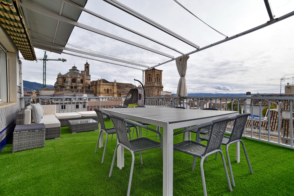 A spacious terrace features a rectangular dining table with several chairs, surrounded by comfortable seating areas. Lush green artificial grass creates a vibrant atmosphere, and views of the cathedral and city skyline are visible in the background under a partly cloudy sky.