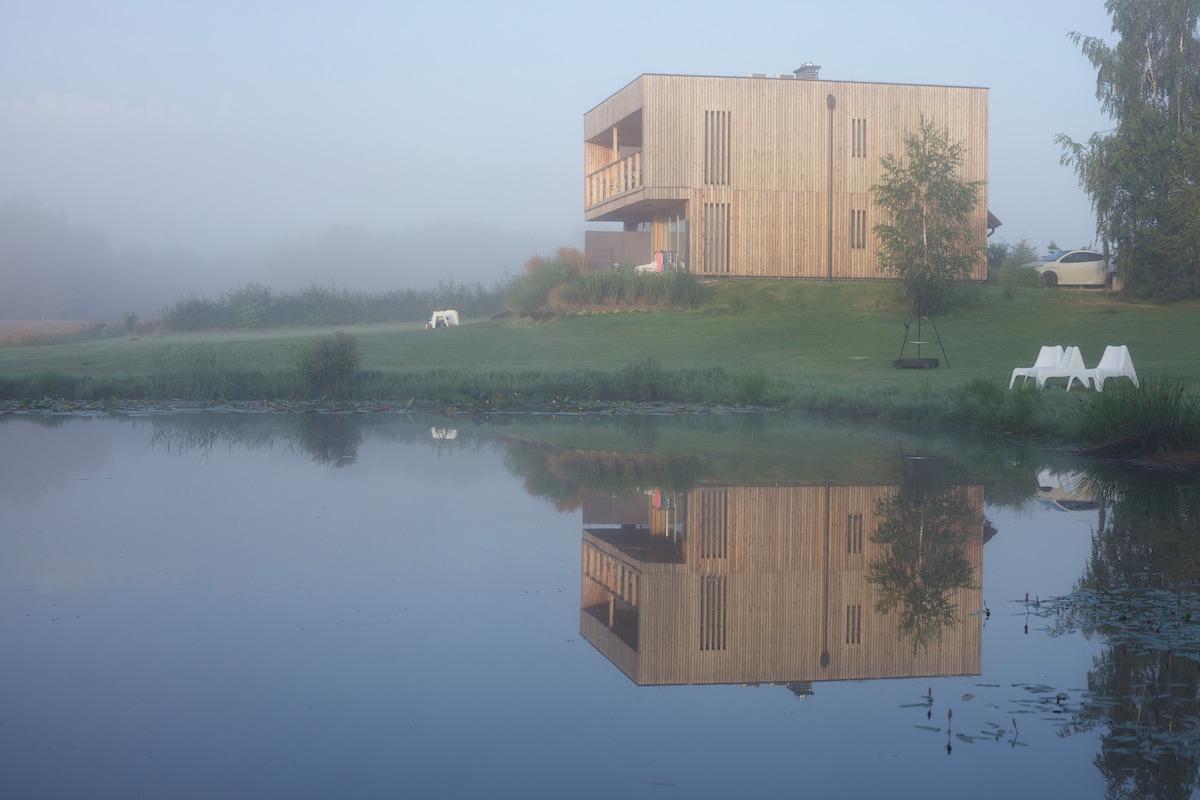 A modern wooden building is reflected in a calm body of water, surrounded by greenery. The structure features an upper balcony, adding to its architectural appeal, while a gentle fog creates a serene atmosphere.