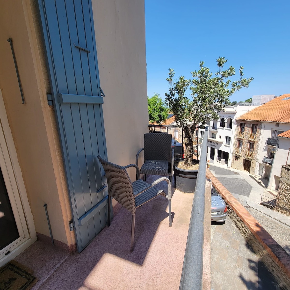 A small balcony area is furnished with two chairs and a table, providing a relaxing outdoor space. A potted olive tree adds greenery, while the surrounding architecture of the village is visible in the background. Bright blue skies create a cheerful atmosphere.