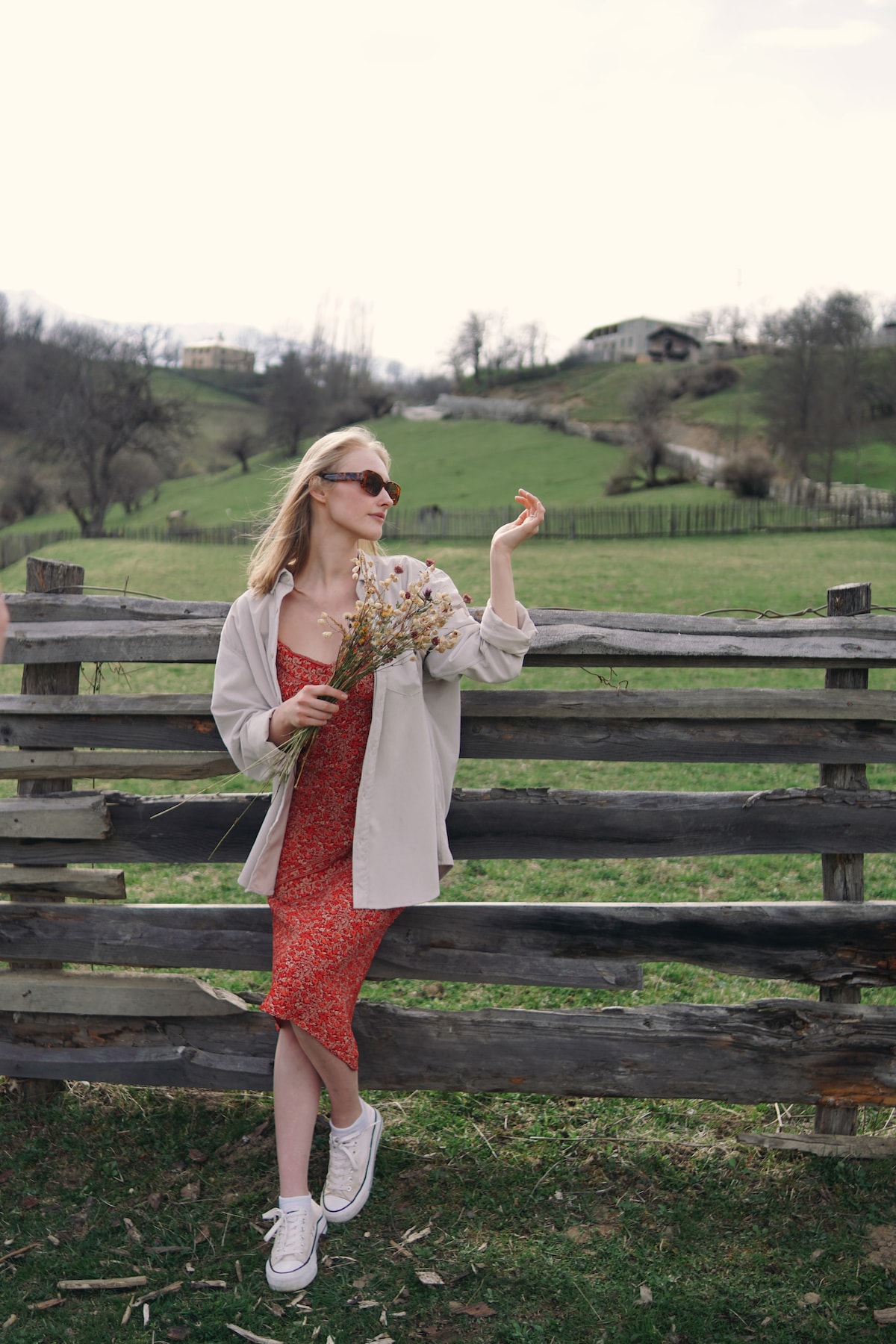 A woman stands gracefully by a rustic wooden fence, holding a bouquet of dried flowers. The lush green landscape extends into the background, featuring rolling hills and a few distant houses, creating a serene and natural setting.