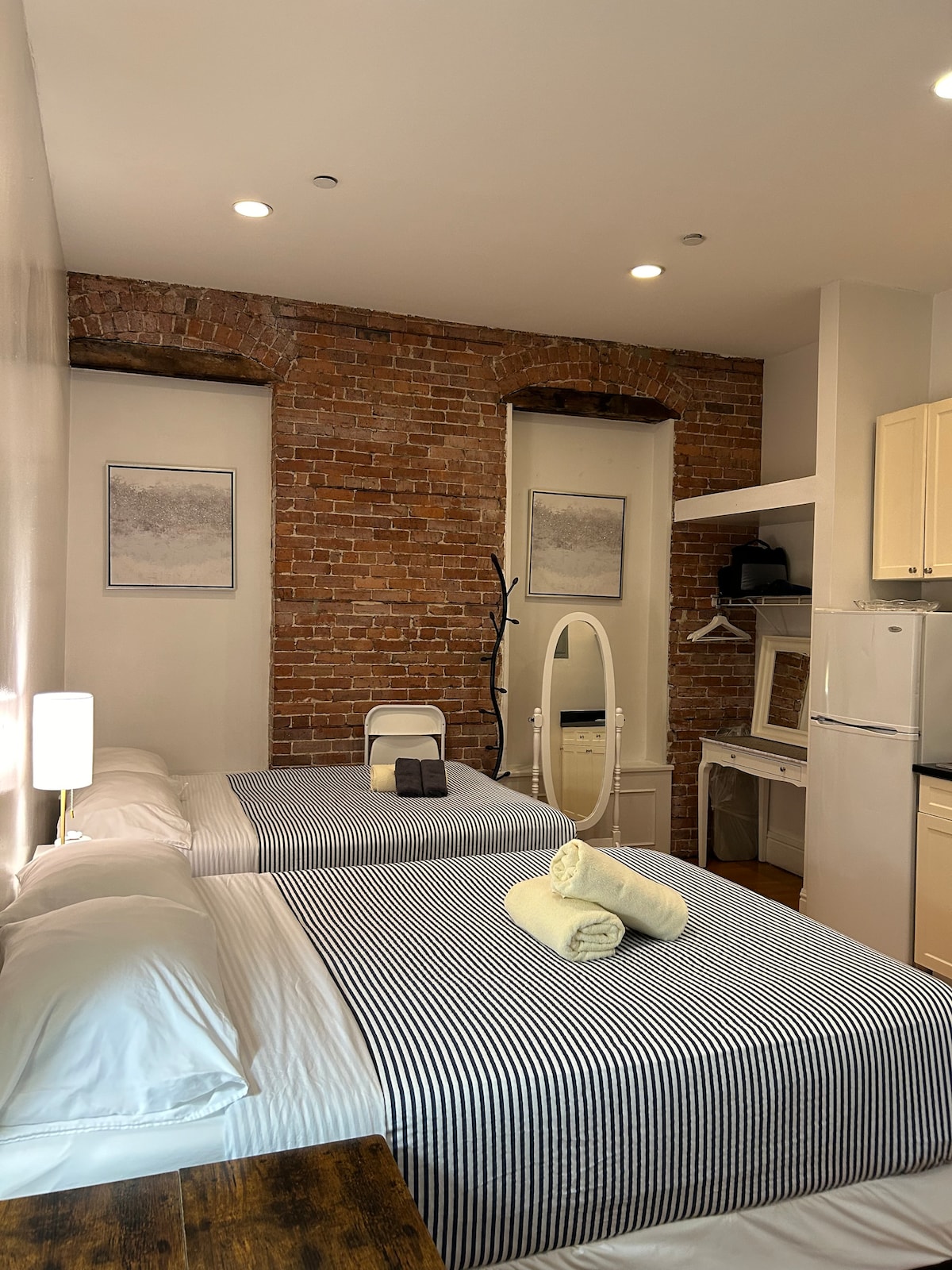 A cozy room showcases two spacious beds, each topped with striped linens. Exposed brick walls add character, while soft lighting highlights a neat arrangement of furniture. A mirror and kitchenette amenities are visible, promoting convenience for guests.