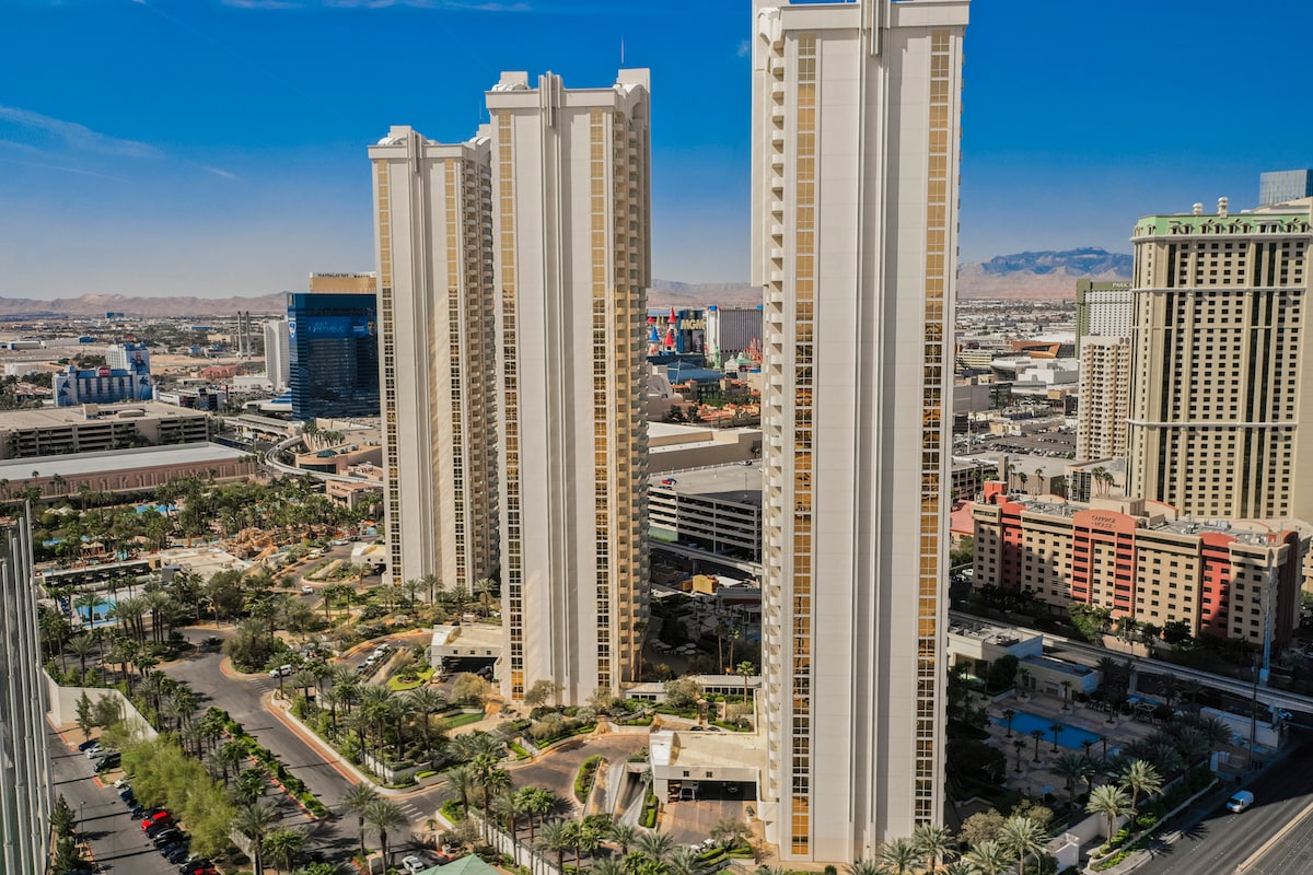 The image showcases three towering high-rise buildings set against a clear blue sky, emphasizing the urban landscape. In the foreground, lush greenery and a pool area are visible, surrounded by palm trees, while the bustling Las Vegas Strip can be seen in the distance.