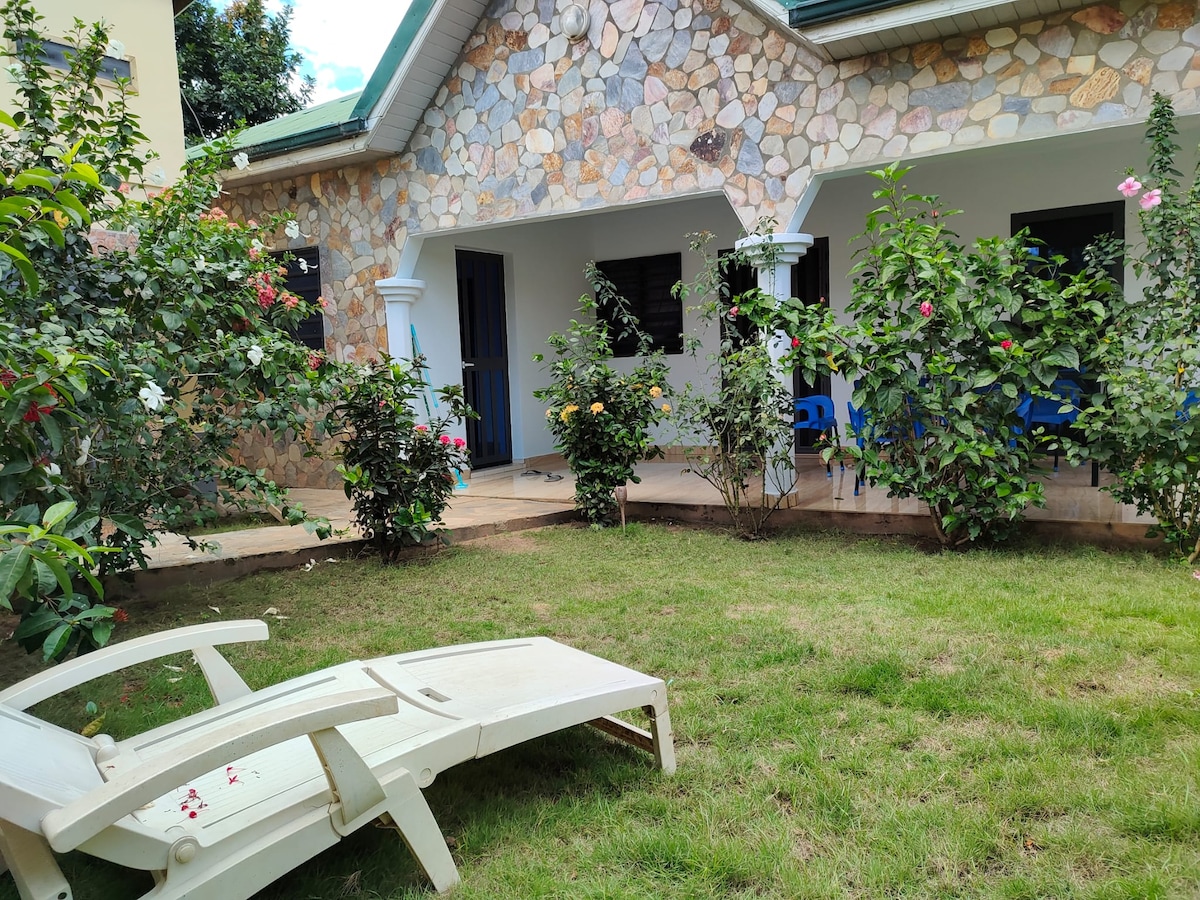 A stone exterior home is framed by a green lawn and flowering shrubs. A white lounge chair is positioned in the foreground, with the entrance and patio area visible. Blue chairs are arranged on the porch, inviting relaxation amidst the well-maintained garden.