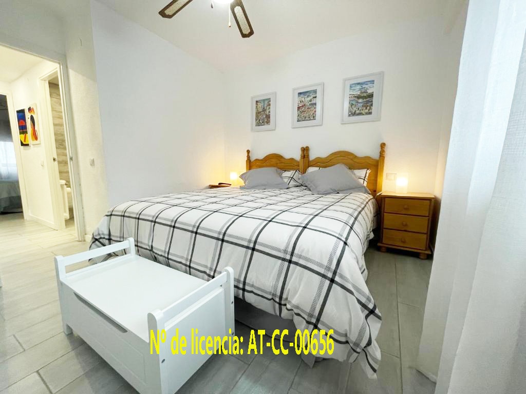 A spacious sleeping area features a large bed with a checkered duvet cover and wooden headboard. Nightstands with lamps are placed on either side of the bed. A small white table is positioned at the foot of the bed, with soft lighting enhancing the calm atmosphere.