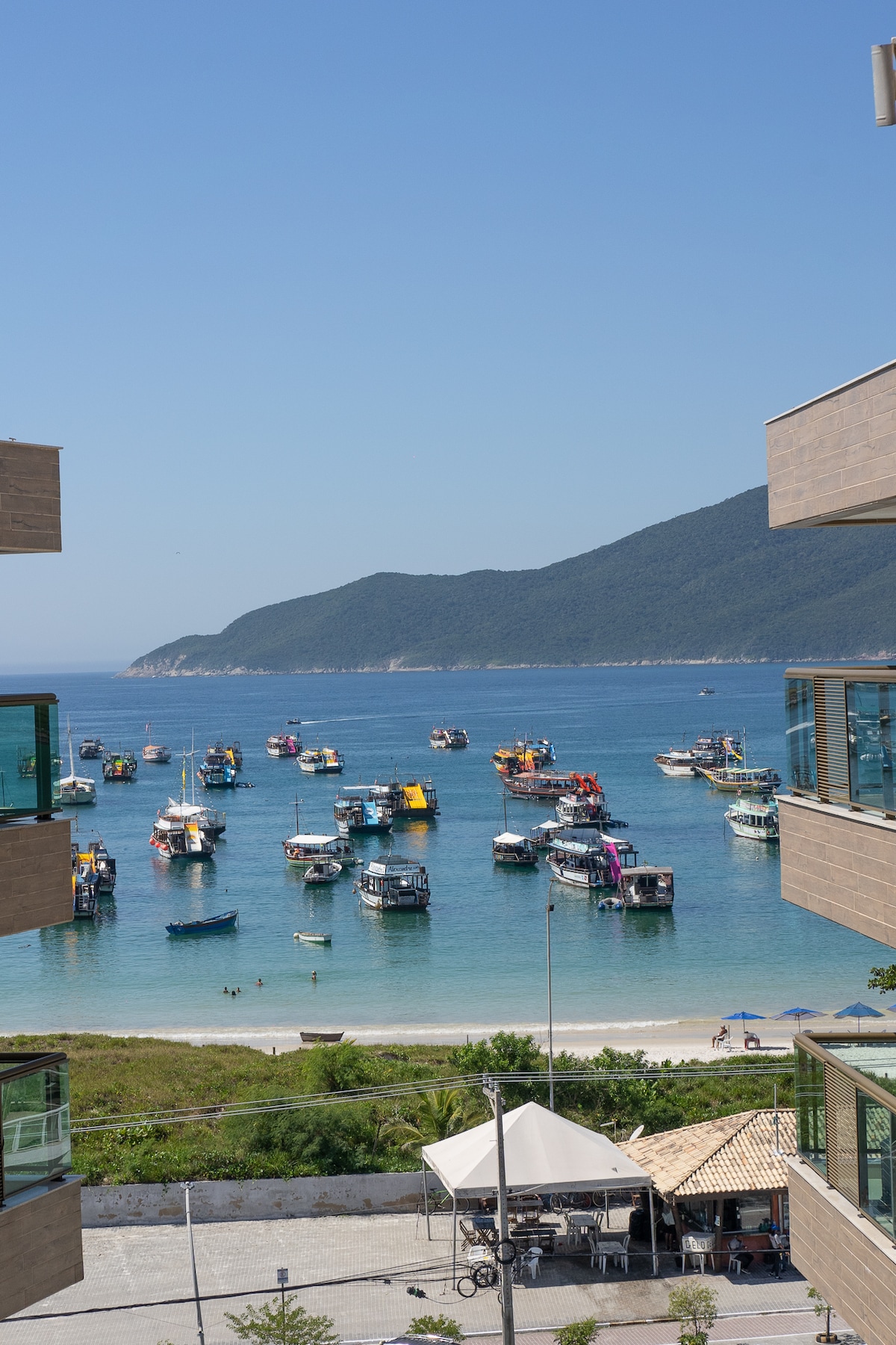 A view of a tranquil bay is presented, filled with colorful fishing boats anchored near the shore. Lush greenery lines the beach, and a gentle slope leads up to a backdrop of mountains under a clear blue sky.