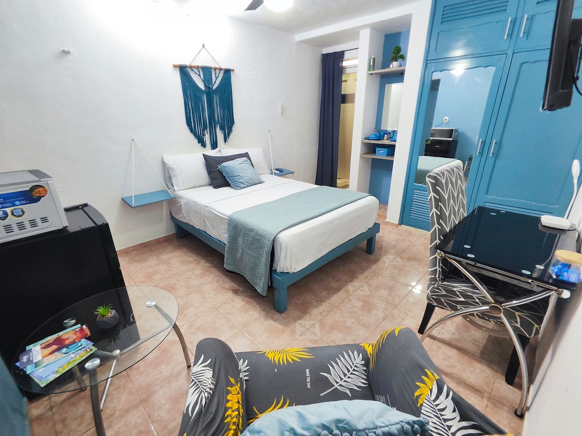 A well-lit room features a comfortable bed with a soft throw blanket and decorative cushions. A sleek desk with a chair and a small glass table are included. The walls are painted in calming blue tones, complemented by storage units and a TV in the corner.