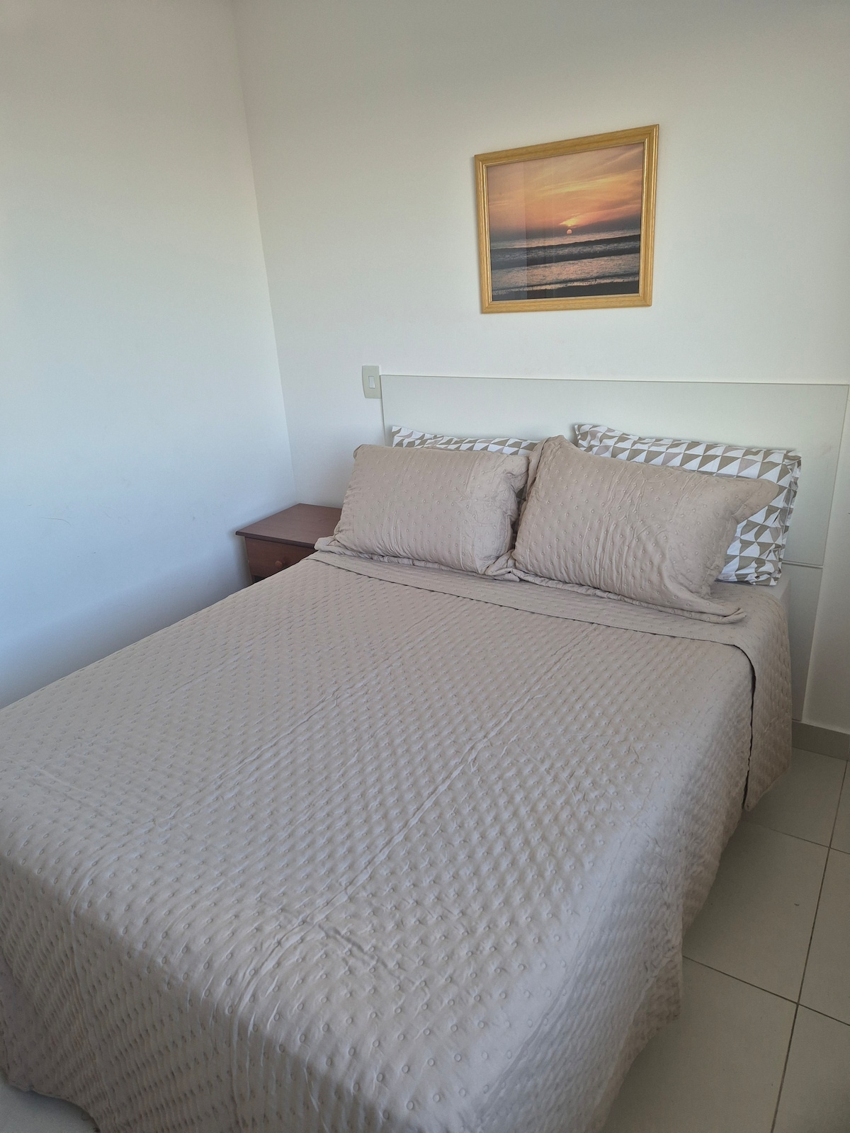 A sleeping area is presented, featuring a spacious bed dressed in light beige bedding, complemented by two matching pillows. A small nightstand is positioned next to the bed, and a framed sunset artwork hangs above, introducing a subtle touch of color to the room.