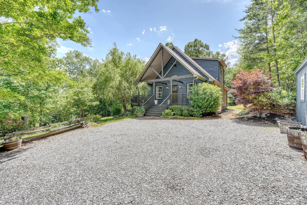 A modern cottage with a steeply pitched roof is surrounded by lush trees and flowering shrubs. A gravel driveway leads to the home, creating an inviting entrance. The landscaped yard features a blend of greenery that complements the cottage's exterior.