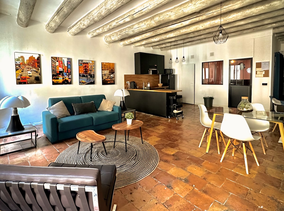 An open living space features exposed wooden beams overhead, with a mix of modern and natural decor. A teal sofa faces two circular side tables placed on a patterned rug. A dining area is positioned nearby, accompanied by a kitchen area with a breakfast bar.