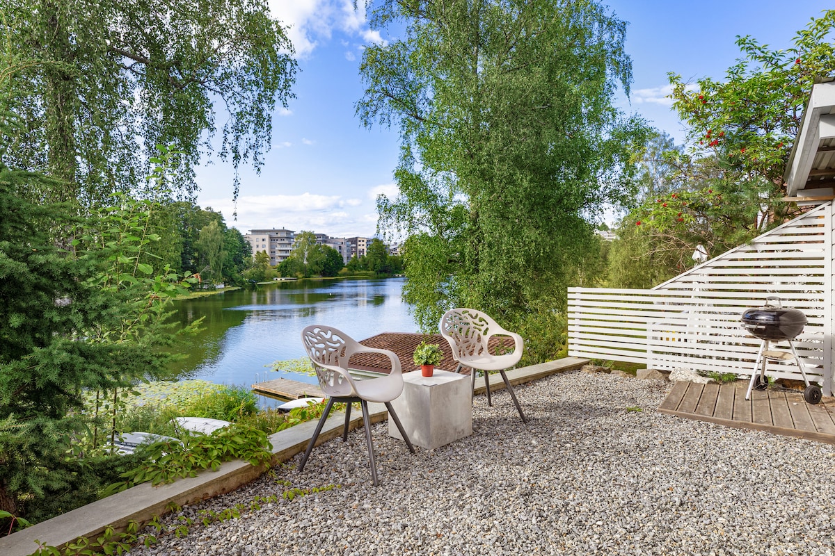 A serene outdoor space is depicted, featuring two white chairs arranged around a round table. The gravel patio overlooks Kolbotnvannet, with lush greenery and reflections on the water creating a peaceful ambience. A grill is positioned nearby, adding to the outdoor experience.