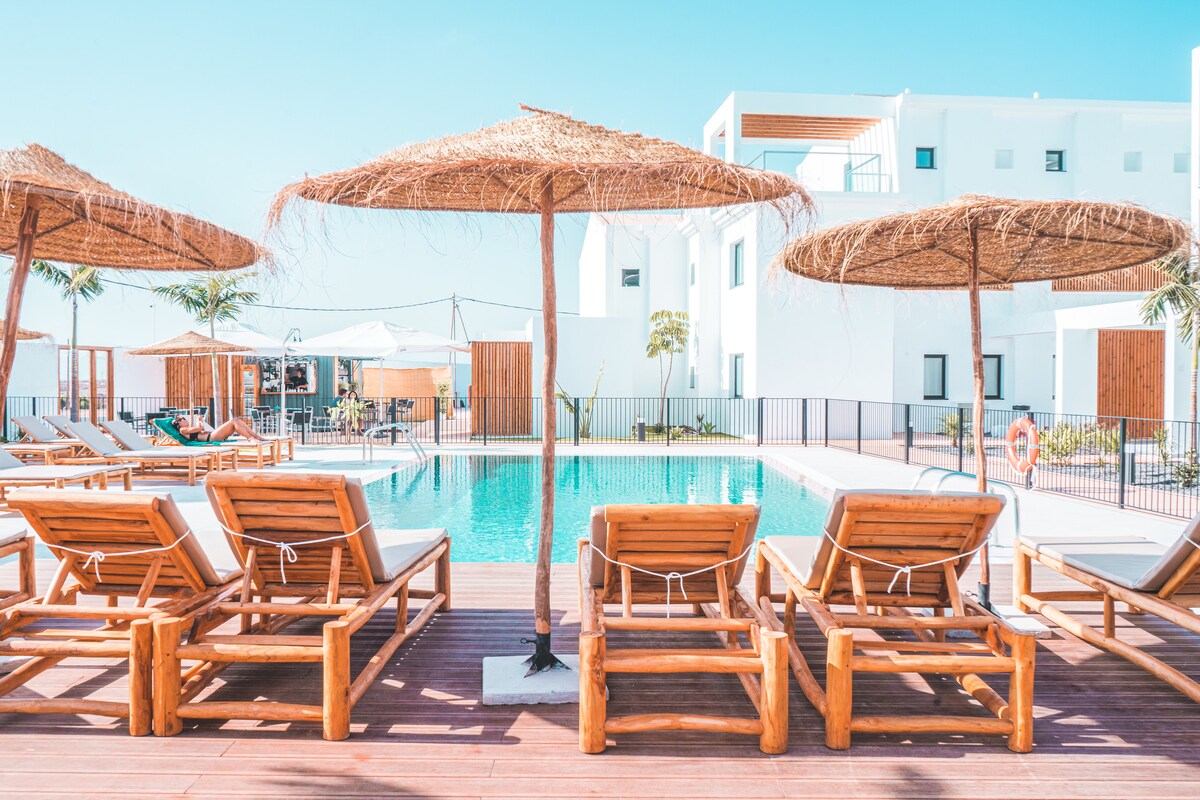 A community pool area is featured, with wooden lounge chairs arranged under thatched umbrellas. The turquoise water of the pool reflects the clear blue sky, surrounded by palm trees and modern white buildings.