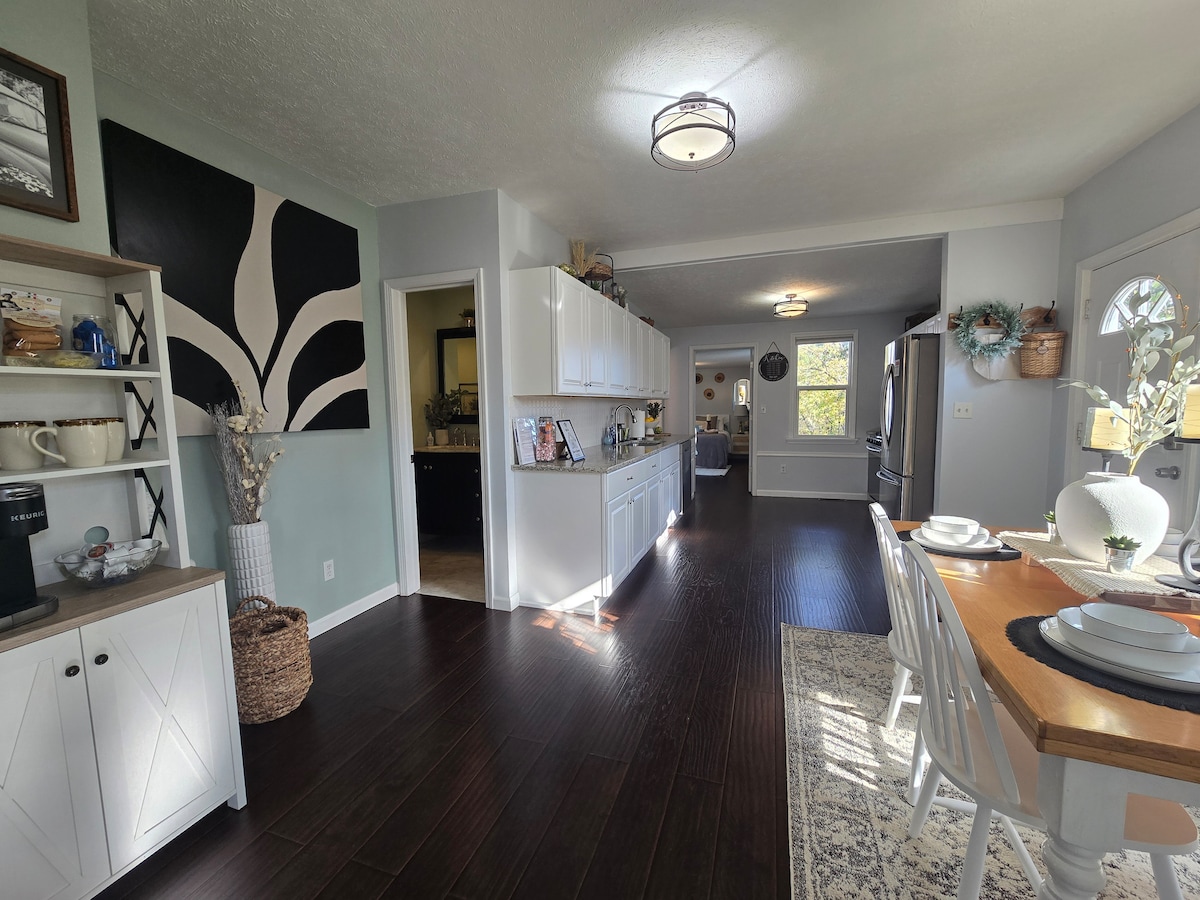 The kitchen and dining area feature an open layout with dark hardwood flooring. White cabinetry contrasts against the muted wall colors. A dining table is set with plates, while natural light brightens the space through a nearby window.