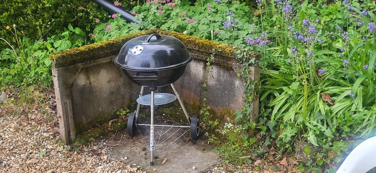 A black charcoal grill is positioned on a stone base, located beside a lush garden with vibrant green foliage and colorful flower blooms. The surrounding area features a gravel pathway, enhancing the outdoor setting.
