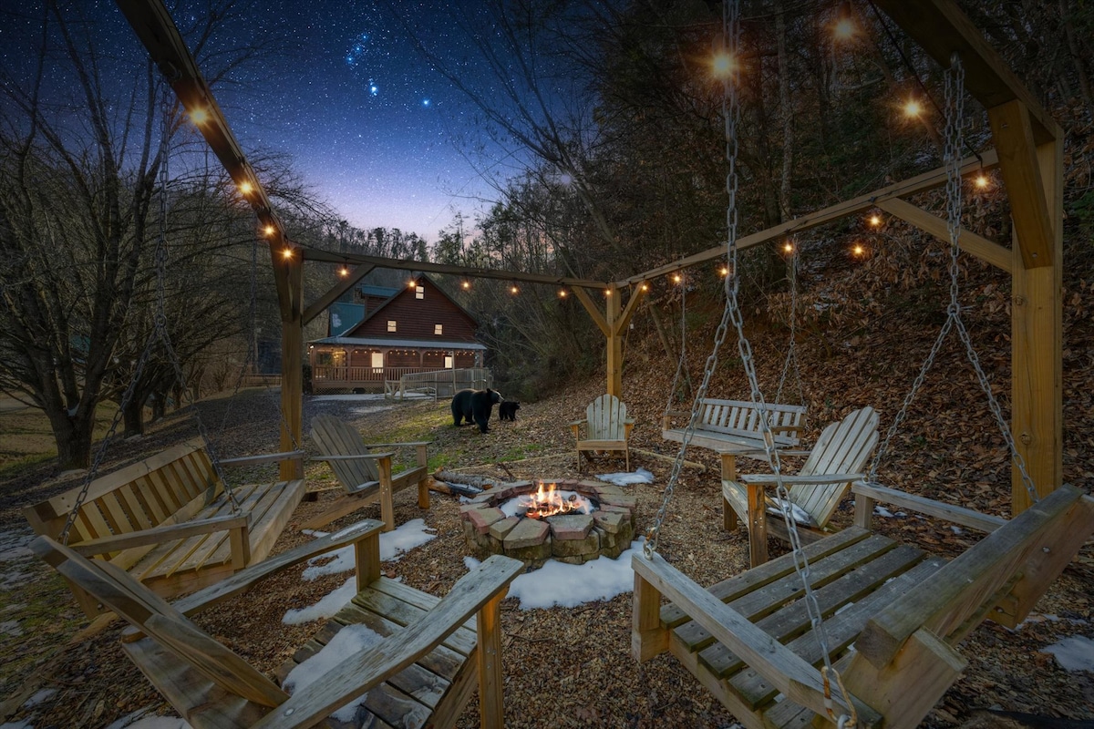 An outdoor fire pit area features multiple wooden chairs arranged around a central fire pit, illuminated by warm string lights. The wooded setting includes a hint of snow on the ground, with gentle lighting from the evening sky and a bear spotted in the background.