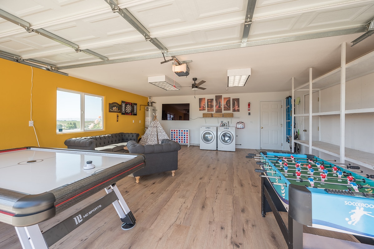 The game room features a bright yellow accent wall, with windows allowing natural light in. An air hockey table and foosball table occupy the space, alongside a comfortable sectional couch. Laundry facilities are visible at the far end, and shelves provide additional storage.