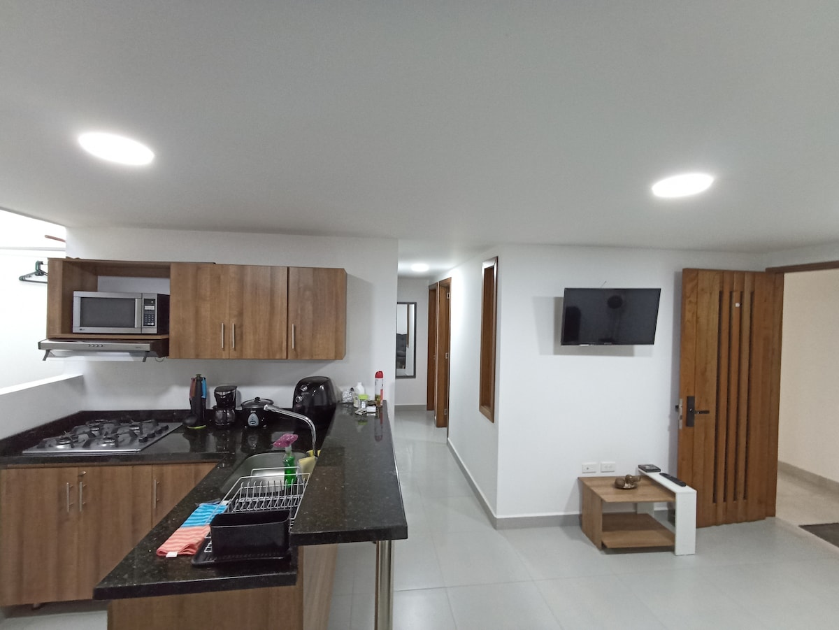 A modern kitchen area showcases sleek wooden cabinetry and a dark countertop. A stove, microwave, and coffee maker are present. Natural light enters through a window, while a television is mounted on the wall opposite. The living space features a neutral color palette and smooth tile flooring.