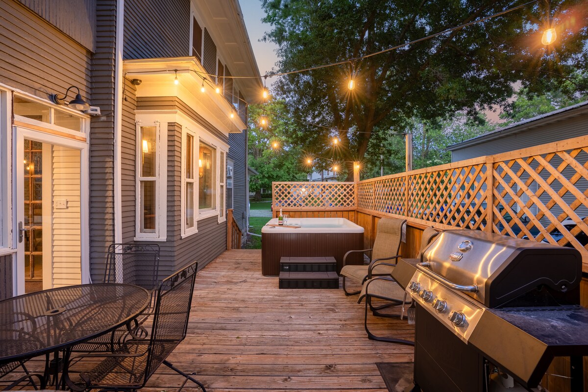 A spacious outdoor deck is illuminated by string lights, featuring a hot tub, a grill, and comfortable seating. A table with chairs is positioned nearby, and a wooden privacy lattice adds a sense of separation to the area, surrounded by greenery.