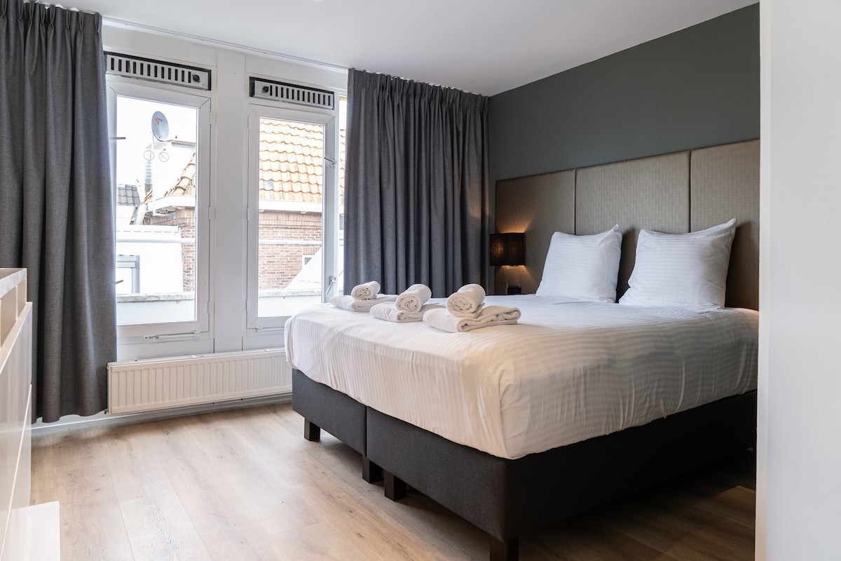 A well-appointed sleeping area features a spacious bed with soft pillows and neatly arranged towels. Natural light enters through two large windows, which are adorned with dark curtains. The light wooden floor enhances the overall brightness of the room.