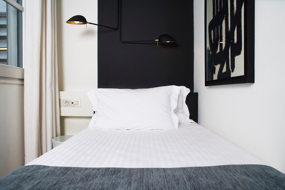 A compact room features a large single bed with crisp white linens and a gray accent blanket. Stylish bedside lighting is attached to the dark wall, and artwork adds visual interest. A window allows natural light to brighten the space, complemented by soft curtains.