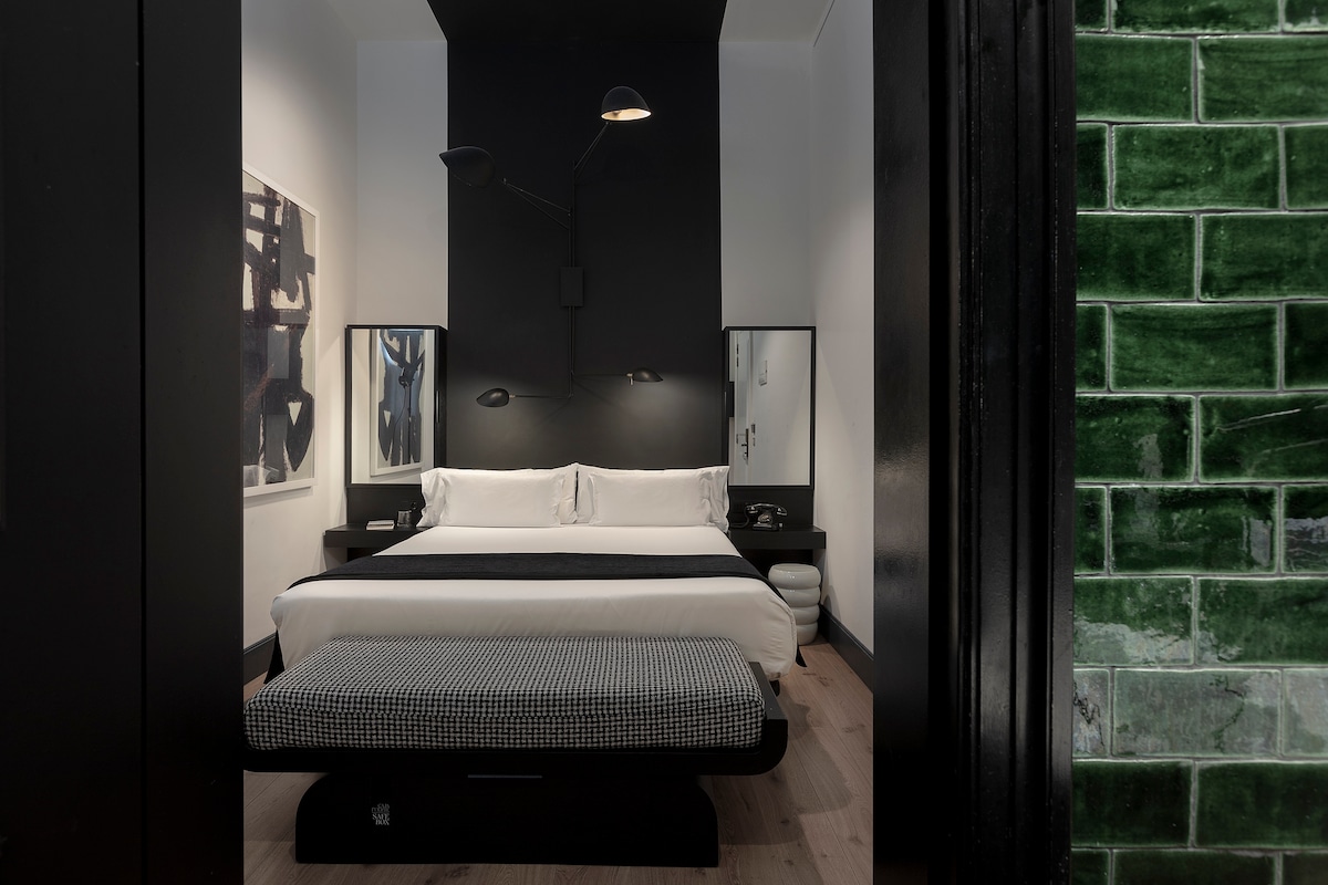 An interior room features a spacious double bed with black accents and white linens. Two mirrors are positioned beside the bed, reflecting minimalistic decor. A modern lamp provides warm lighting. Textured green tiles are visible in the adjacent space, creating an interesting design contrast.