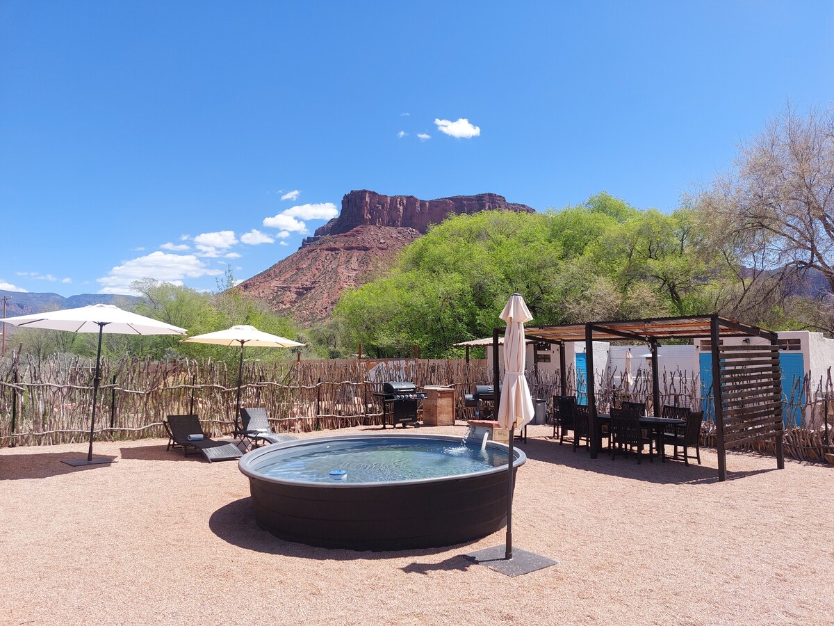A spacious outdoor area features a round cowboy pool at the center, surrounded by lounge chairs, umbrellas, and a shaded dining space. A rustic wooden fence encloses the setting, with desert vegetation and a striking rock formation in the background under a clear blue sky.