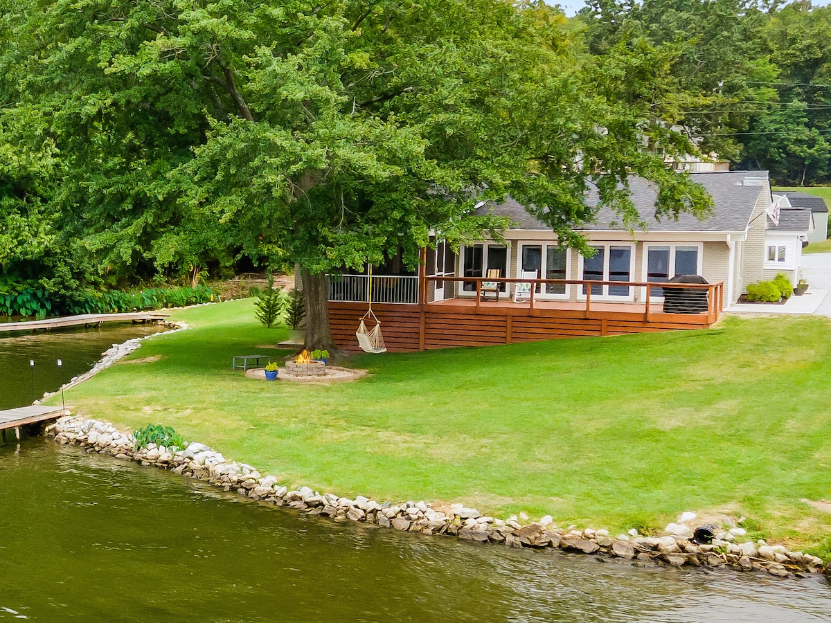 A lakeside home is nestled within a gently sloped yard. A large tree offers shade beside a wooden deck featuring a screened porch. A stone fire pit and seating area are visible on the lush green lawn, leading to a rippling lake with a floating dock.