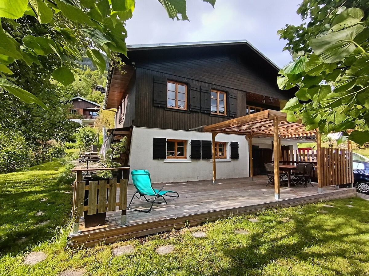 The exterior of a two-story chalet is depicted, featuring a dark wood facade with black window shutters. A sunny terrace is visible, accompanied by a green lounge chair and a wooden dining table. Lush greenery surrounds the chalet, enhancing the tranquil mountain setting.