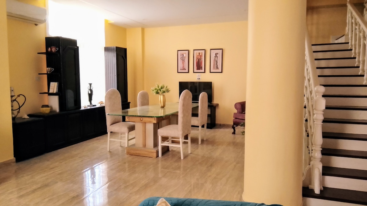 A spacious living area is highlighted by a large dining table surrounded by six upholstered chairs. Soft yellow walls complement the tiled floor, while three art pieces are displayed on the wall. Natural light streams in through the large window, enhancing the open atmosphere.