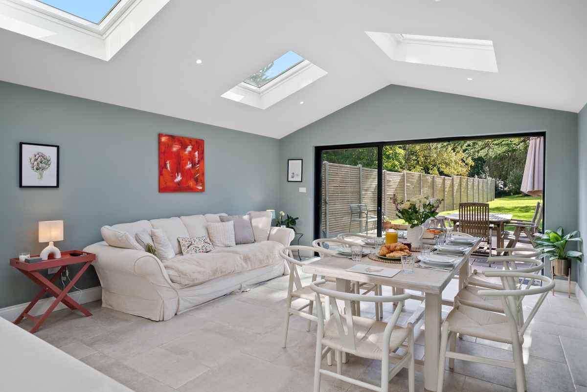 A bright and airy living area features large windows and skylights, allowing natural light to fill the space. A comfortable sofa and a dining table with white chairs provide inviting seating options. Views of the garden can be seen through sliding glass doors.