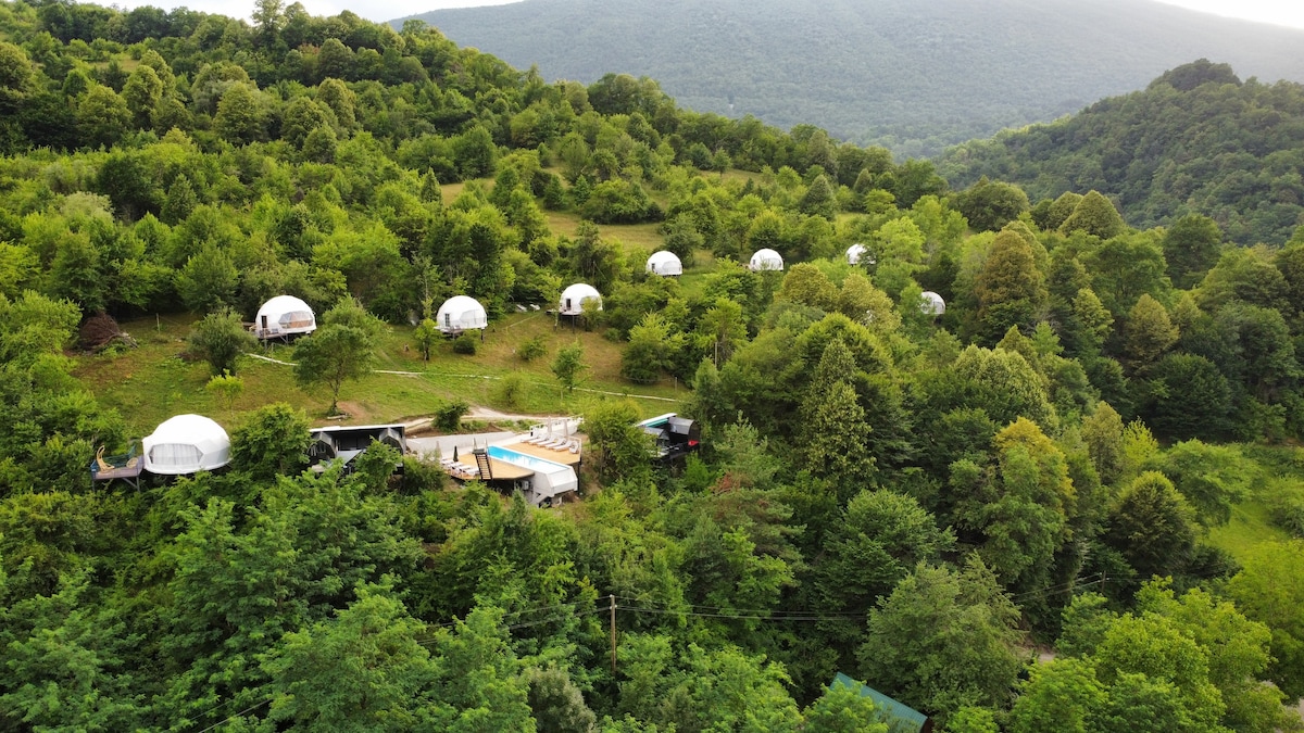 Aerial view of a glamping site nestled in lush green hills. Several luxury tents are positioned among trees, featuring a communal area with a swimming pool. The surrounding landscape offers a blend of nature and outdoor accommodation in a serene mountainous setting.
