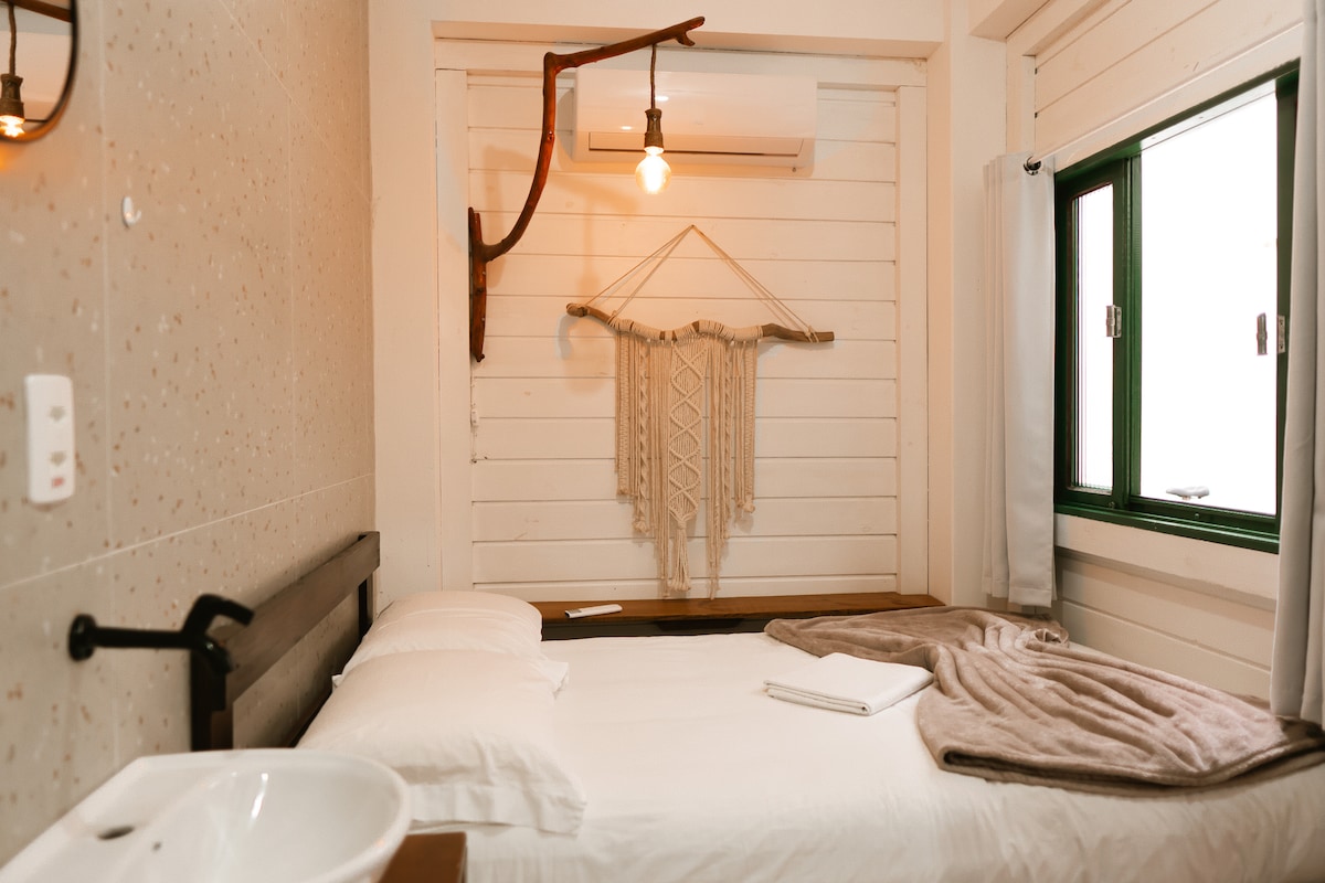 A cozy sleeping area features a spacious bed adorned with neutral-toned linens and a soft blanket. Natural light enters through a green-framed window, while a decorative wall piece adds texture. An air conditioning unit and a wall-mounted light provide comfort and functionality.