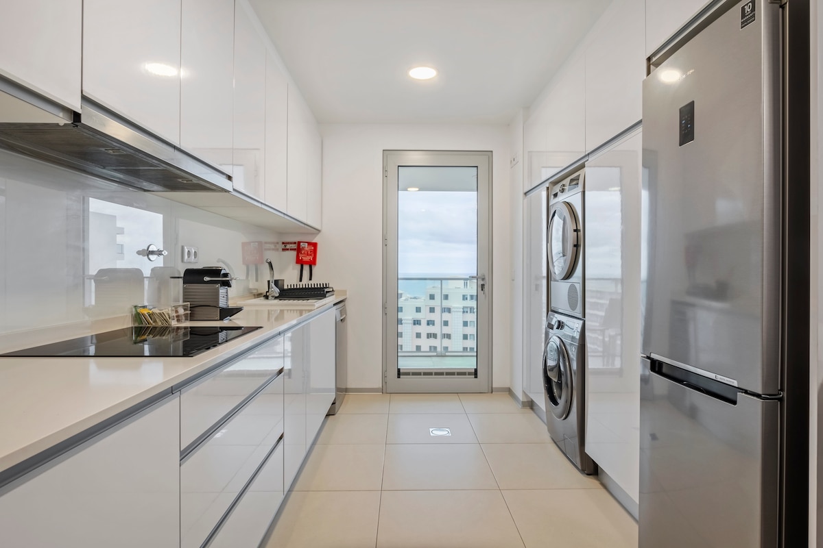 A modern kitchen features sleek white cabinetry and stainless steel appliances, including a refrigerator and washer/dryer. A window offers a view of the ocean, allowing natural light to fill the space. Clean lines and a minimalist design create an inviting atmosphere.