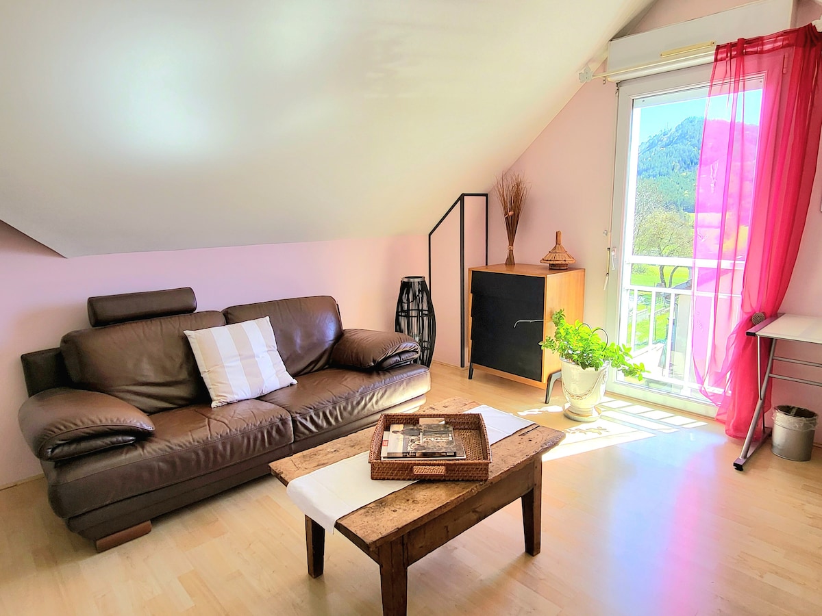 A light-filled living area features a brown leather sofa accompanied by a white cushion. A wooden coffee table sits centrally, topped with decorative items. A tall black cabinet stands against one wall. A glass door leads to a balcony, adorned with flowing sheer curtains and a view of surrounding greenery.